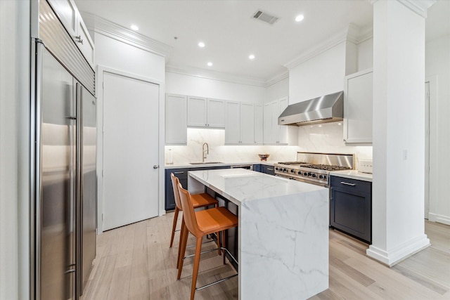 kitchen with built in refrigerator, wall chimney exhaust hood, a sink, ornamental molding, and light wood-type flooring