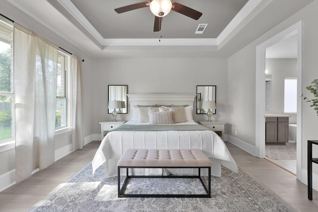 bedroom with a tray ceiling, light wood-style floors, crown molding, ensuite bathroom, and baseboards
