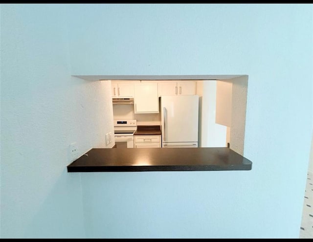 kitchen view of white appliances, a peninsula, under cabinet range hood, dark countertops, and white cabinets