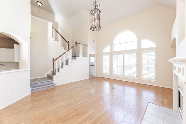 unfurnished living room with high vaulted ceiling, stairs, light wood finished floors, a fireplace with flush hearth, and a chandelier