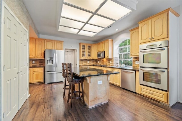 kitchen featuring appliances with stainless steel finishes, light brown cabinetry, glass insert cabinets, a kitchen breakfast bar, and dark wood finished floors