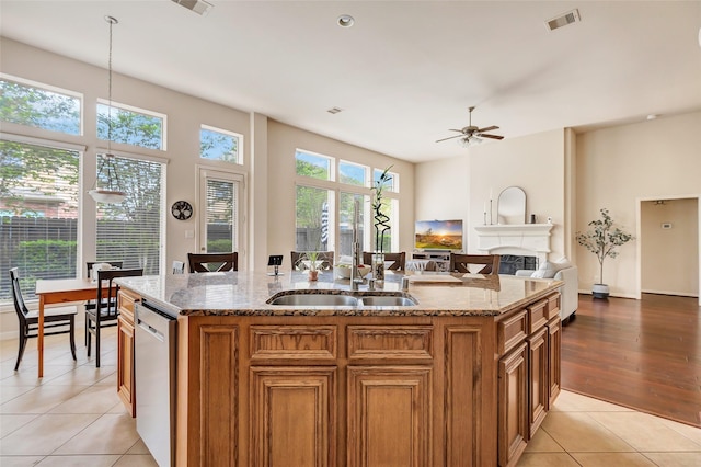 kitchen featuring brown cabinets, dishwasher, a kitchen island with sink, light tile patterned floors, and light stone countertops