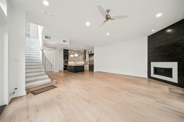 unfurnished living room with ceiling fan, light wood-style floors, a large fireplace, stairway, and recessed lighting