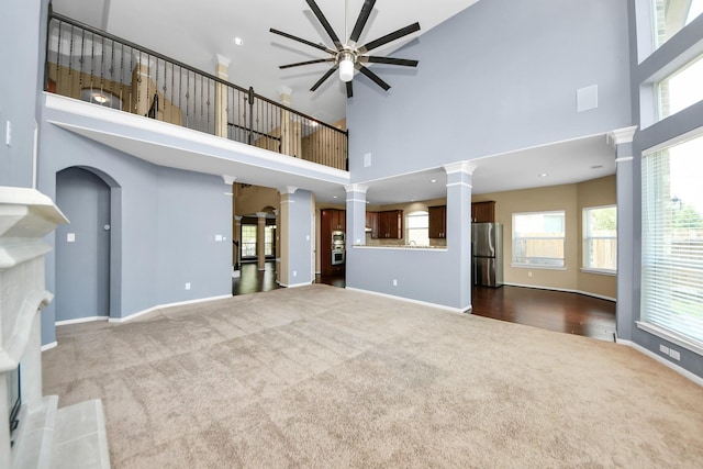 unfurnished living room with dark carpet, ornate columns, ceiling fan, a high ceiling, and plenty of natural light