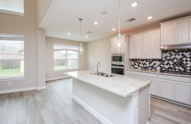 kitchen with a sink, stainless steel appliances, a center island with sink, backsplash, and light wood-style floors