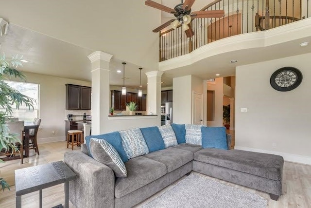 living room with a ceiling fan, ornate columns, baseboards, light wood-style flooring, and a towering ceiling