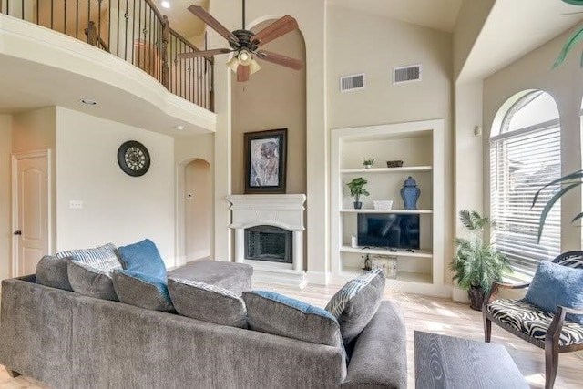 living area with built in shelves, arched walkways, a ceiling fan, high vaulted ceiling, and a fireplace with raised hearth