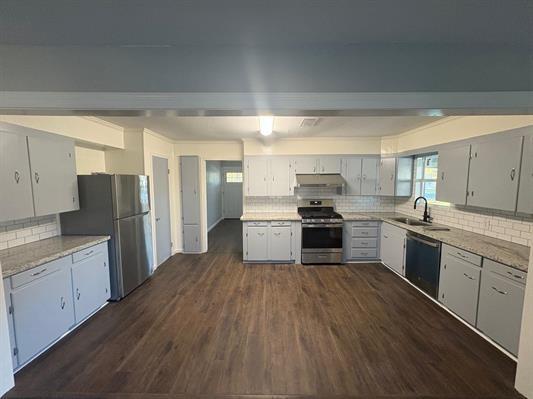 6240 25th St, Groves, TX 77619 - kitchen with appliances with stainless steel finishes, decorative backsplash, under cabinet range hood, dark wood-style flooring, and a sink