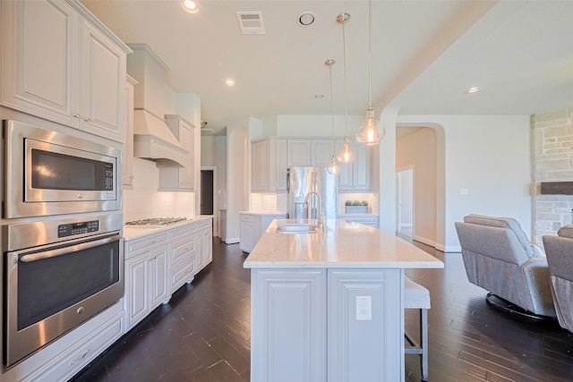 kitchen with appliances with stainless steel finishes, open floor plan, dark wood-style flooring, arched walkways, and a kitchen island with sink