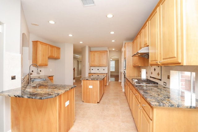 kitchen with a sink, dark stone counters, light brown cabinets, stainless steel gas cooktop, and recessed lighting