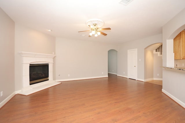 unfurnished living room with a ceiling fan, arched walkways, wood finished floors, and a glass covered fireplace