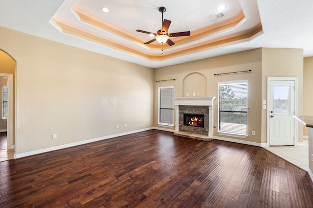unfurnished living room featuring a tray ceiling, ceiling fan, arched walkways, wood-type flooring, and a fireplace