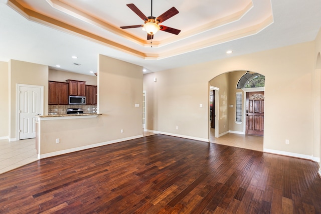 unfurnished living room with arched walkways, a raised ceiling, a ceiling fan, wood finished floors, and recessed lighting