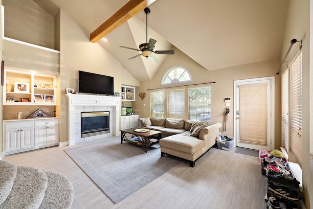 living room featuring high vaulted ceiling, light wood finished floors, a tile fireplace, beam ceiling, and ceiling fan