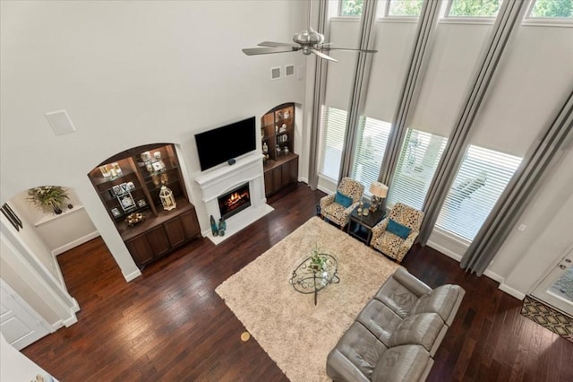 living room featuring ceiling fan, dark wood-style floors, a warm lit fireplace, baseboards, and a high ceiling
