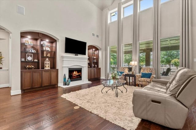 living area featuring dark wood finished floors, a lit fireplace, a towering ceiling, baseboards, and built in features