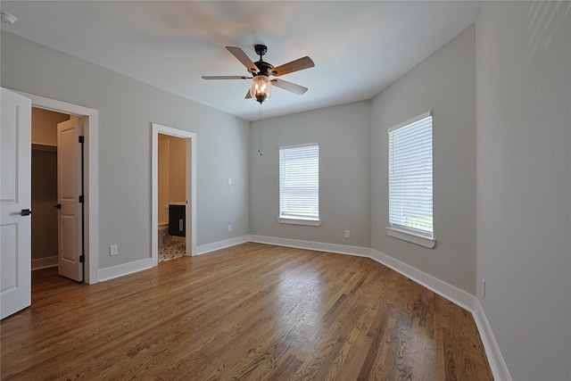 unfurnished bedroom featuring wood finished floors, a walk in closet, baseboards, connected bathroom, and ceiling fan