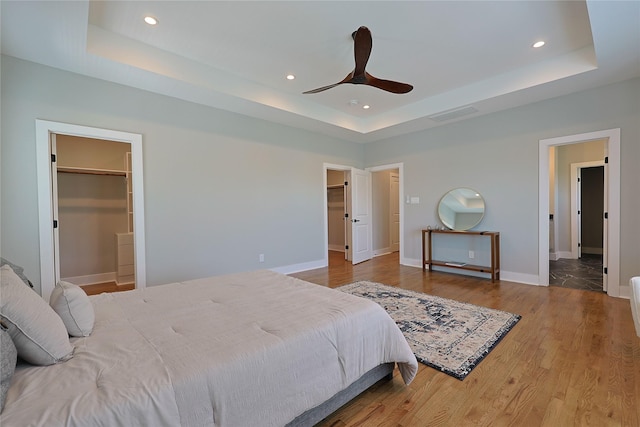bedroom with a raised ceiling, recessed lighting, a walk in closet, wood finished floors, and baseboards