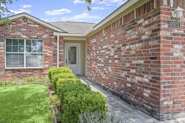view of exterior entry featuring brick siding, roof with shingles, and a yard