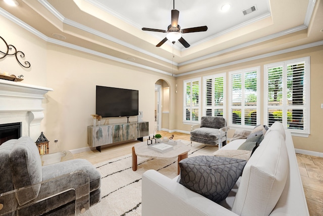 living area featuring a raised ceiling, crown molding, arched walkways, a ceiling fan, and baseboards