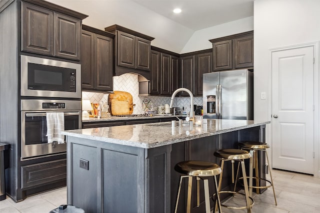 kitchen with stainless steel appliances, a sink, light stone counters, backsplash, and an island with sink