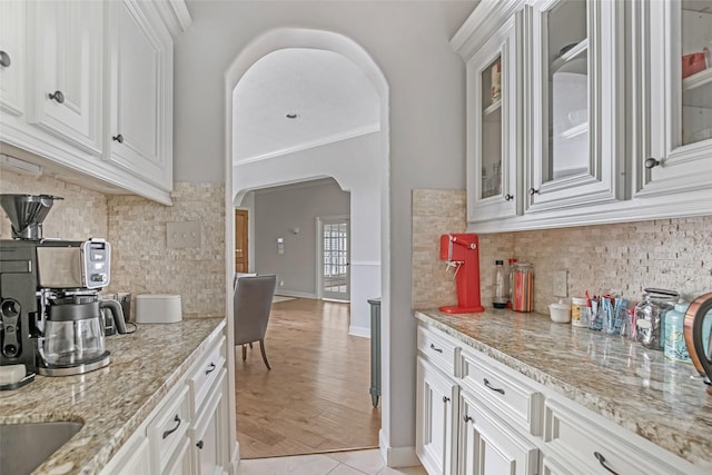 kitchen featuring arched walkways, tasteful backsplash, glass insert cabinets, light stone counters, and crown molding
