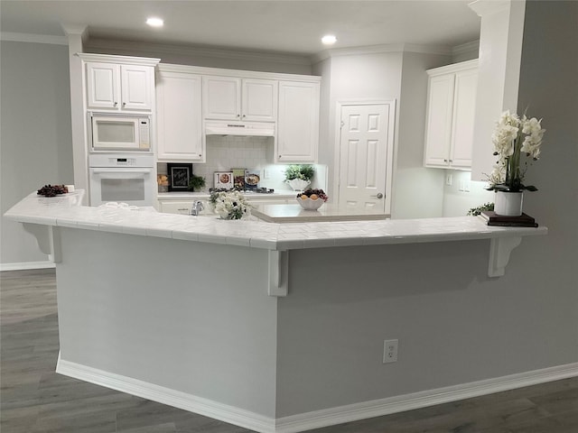kitchen featuring white appliances, under cabinet range hood, tile countertops, baseboards, and white cabinetry