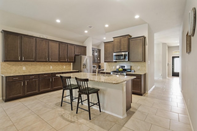 22078 Sun Haven Ln, Porter, TX 77365 - kitchen featuring appliances with stainless steel finishes, light tile patterned flooring, tasteful backsplash, an island with sink, and dark brown cabinetry