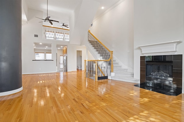 unfurnished living room featuring wood-type flooring, a ceiling fan, a high ceiling, a tile fireplace, and stairs