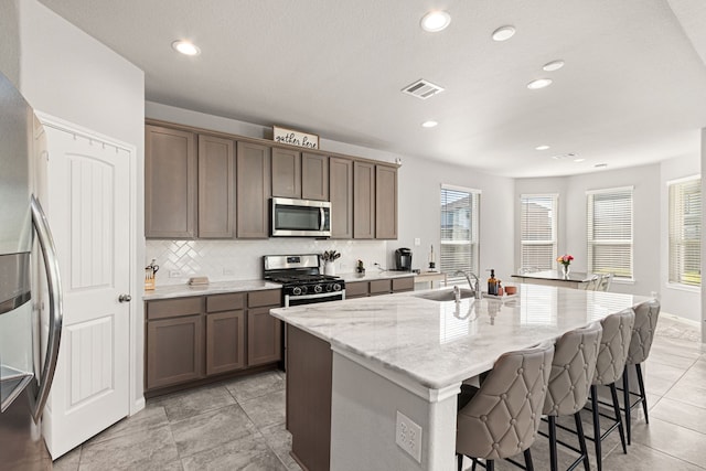 kitchen featuring stainless steel appliances, a sink, an island with sink, light stone counters, and a breakfast bar