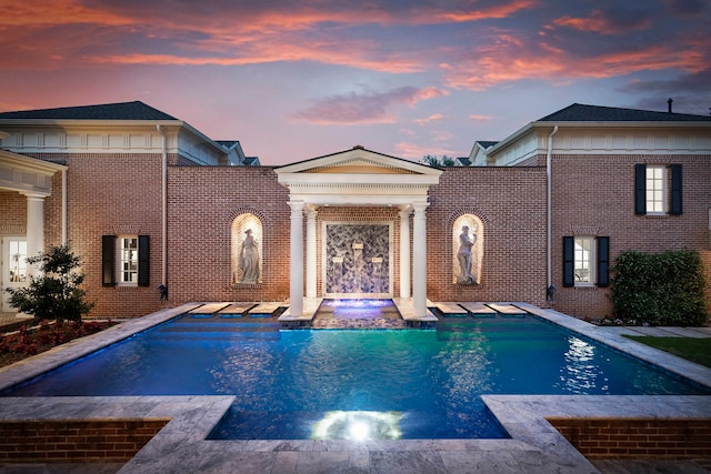 back of property at dusk with brick siding and a pool with connected hot tub