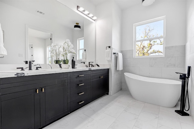 bathroom featuring marble finish floors, tile walls, a freestanding tub, double vanity, and a wainscoted wall