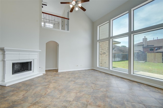 unfurnished living room with ceiling fan, arched walkways, high vaulted ceiling, baseboards, and a fireplace with raised hearth