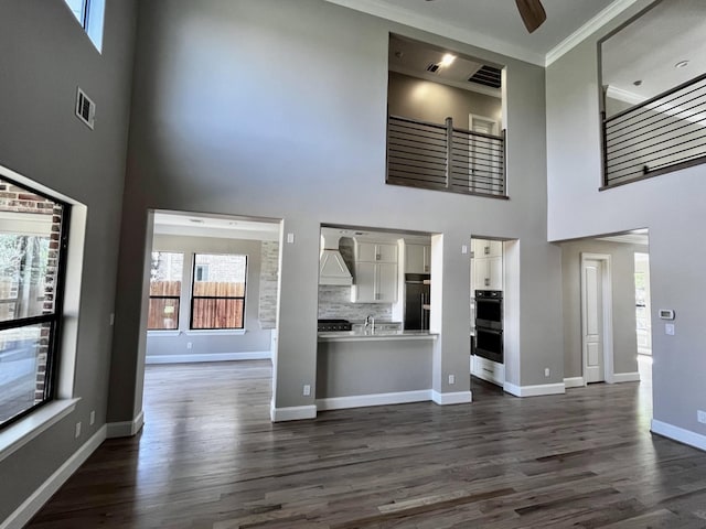 unfurnished living room featuring a high ceiling, ceiling fan, dark wood-style flooring, crown molding, and baseboards