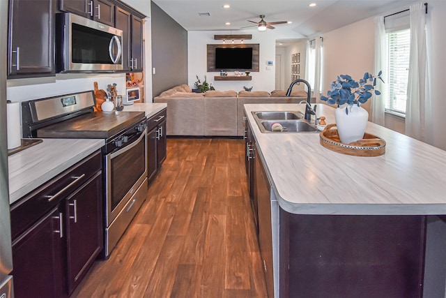 kitchen featuring stainless steel appliances, a sink, a kitchen island with sink, dark wood-type flooring, and a ceiling fan