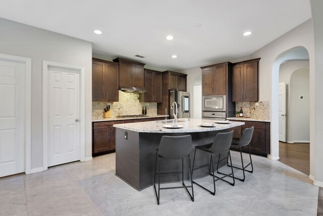6706 Miller Shadow Ln, Sugar Land, TX 77479 - kitchen featuring appliances with stainless steel finishes, a breakfast bar, arched walkways, under cabinet range hood, and decorative backsplash