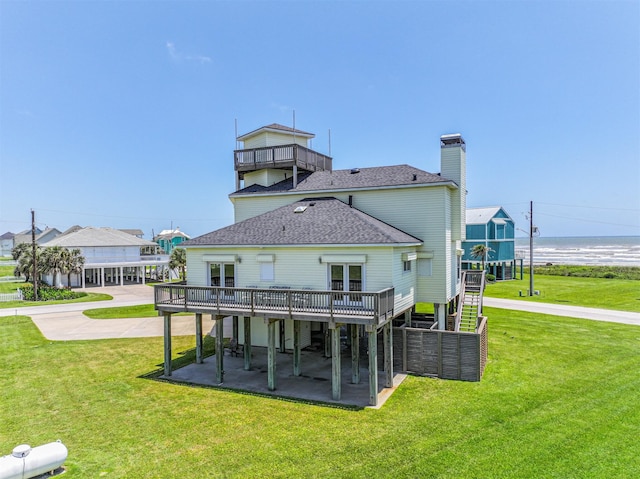 back of house with a shingled roof, a deck with water view, a patio area, a chimney, and a lawn