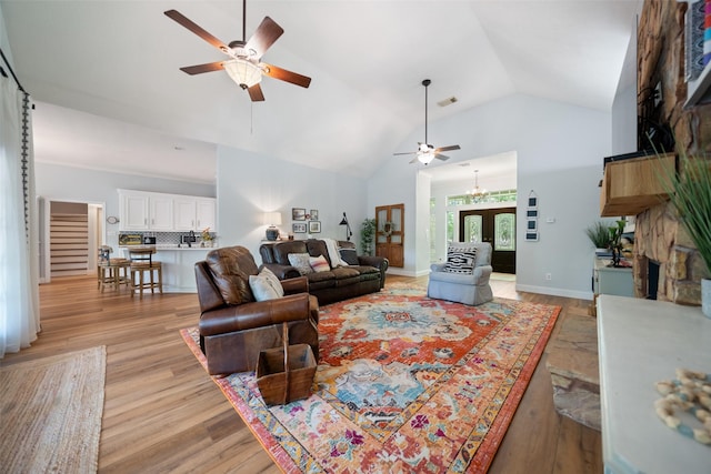 living room with ceiling fan, stairway, light wood-style floors, a stone fireplace, and high vaulted ceiling