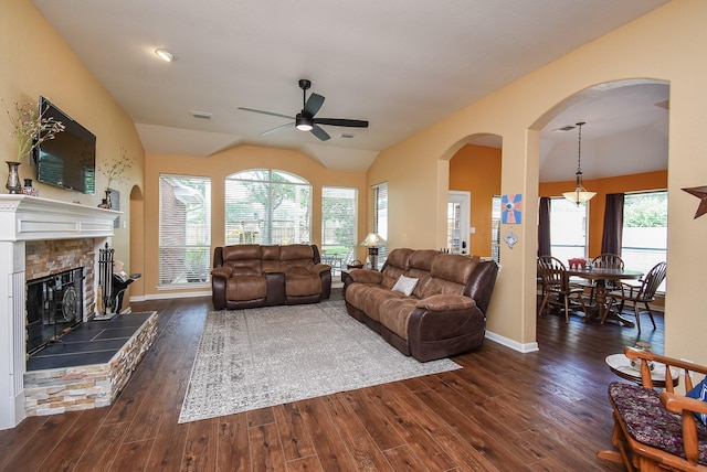 living area with arched walkways, ceiling fan, dark wood-style floors, vaulted ceiling, and a stone fireplace