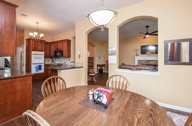 dining area featuring dark wood-type flooring, ceiling fan, arched walkways, a chandelier, and a stone fireplace