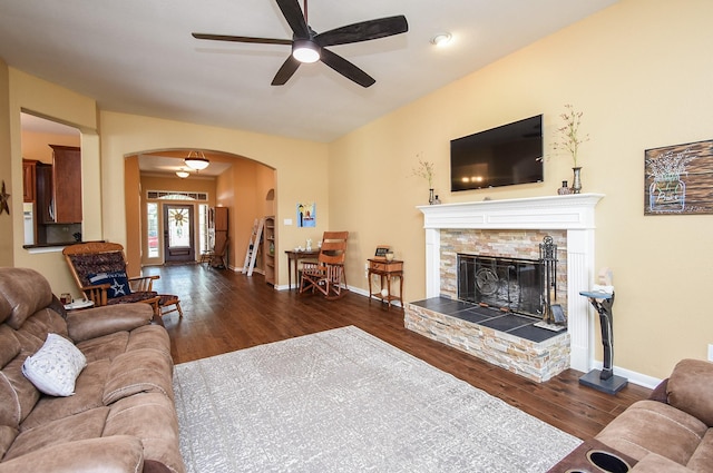 living area with arched walkways, wood finished floors, ceiling fan, a stone fireplace, and baseboards