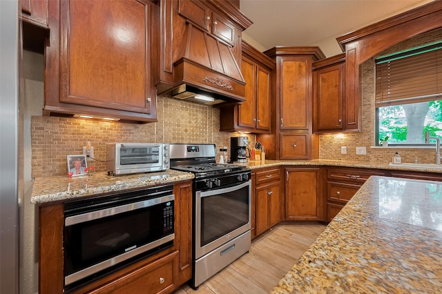 14844 Pollux Dr, Willis, TX 77318 - kitchen featuring stainless steel appliances, premium range hood, light wood-type flooring, a sink, and brown cabinetry