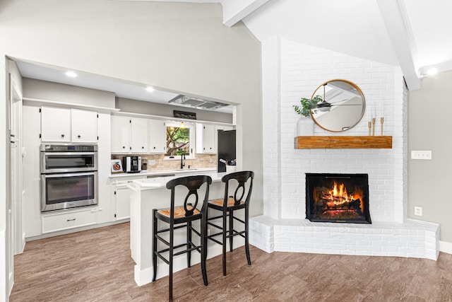 kitchen with a sink, light wood finished floors, beamed ceiling, a brick fireplace, and white cabinets