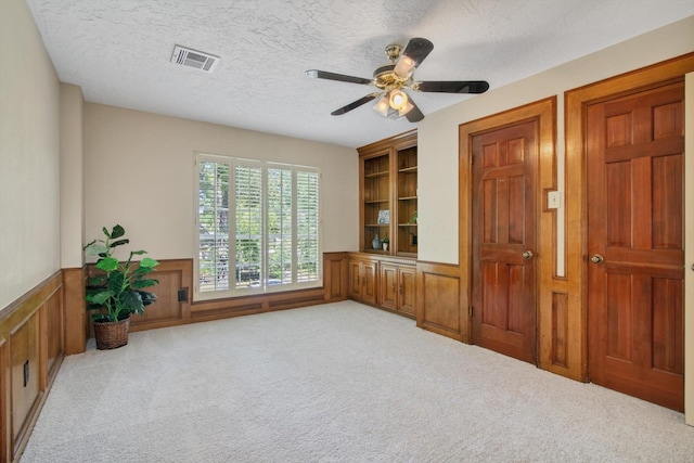 unfurnished room featuring built in features, wainscoting, carpet, a textured ceiling, and a ceiling fan