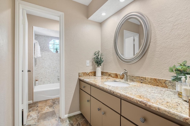 full bath with a textured wall, vanity, stone tile flooring, and baseboards