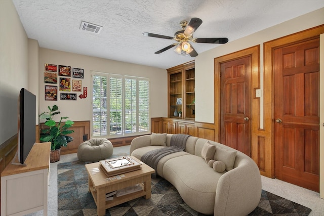 living room featuring carpet, ceiling fan, a textured ceiling, built in shelves, and a wainscoted wall