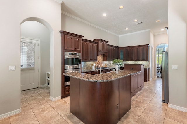 15557 Crown Oaks Dr, Montgomery, TX 77316 - kitchen featuring arched walkways, stainless steel appliances, an island with sink, backsplash, and light tile patterned floors