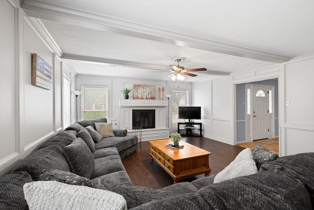 living area with a decorative wall, wood finished floors, crown molding, a glass covered fireplace, and ceiling fan