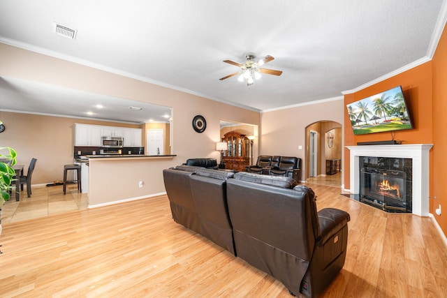 living area with arched walkways, ceiling fan, light wood-style flooring, crown molding, and a high end fireplace