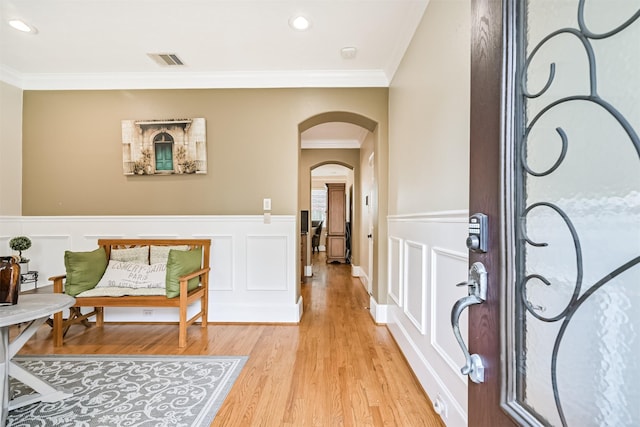 foyer entrance featuring ornamental molding, arched walkways, light wood-style floors, a wainscoted wall, and a decorative wall